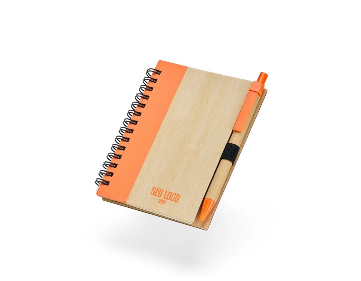 Caderno com capa de casca de bambu e faixa colorida, encadernação Wire-O e cerca de 70 folhas pautadas amareladas. Acompanha caneta esferográfica azul (0,8 mm), com corpo de papelão, clip, ponteira plástica e acionamento por clique. Elástico na capa para fixar a caneta.
